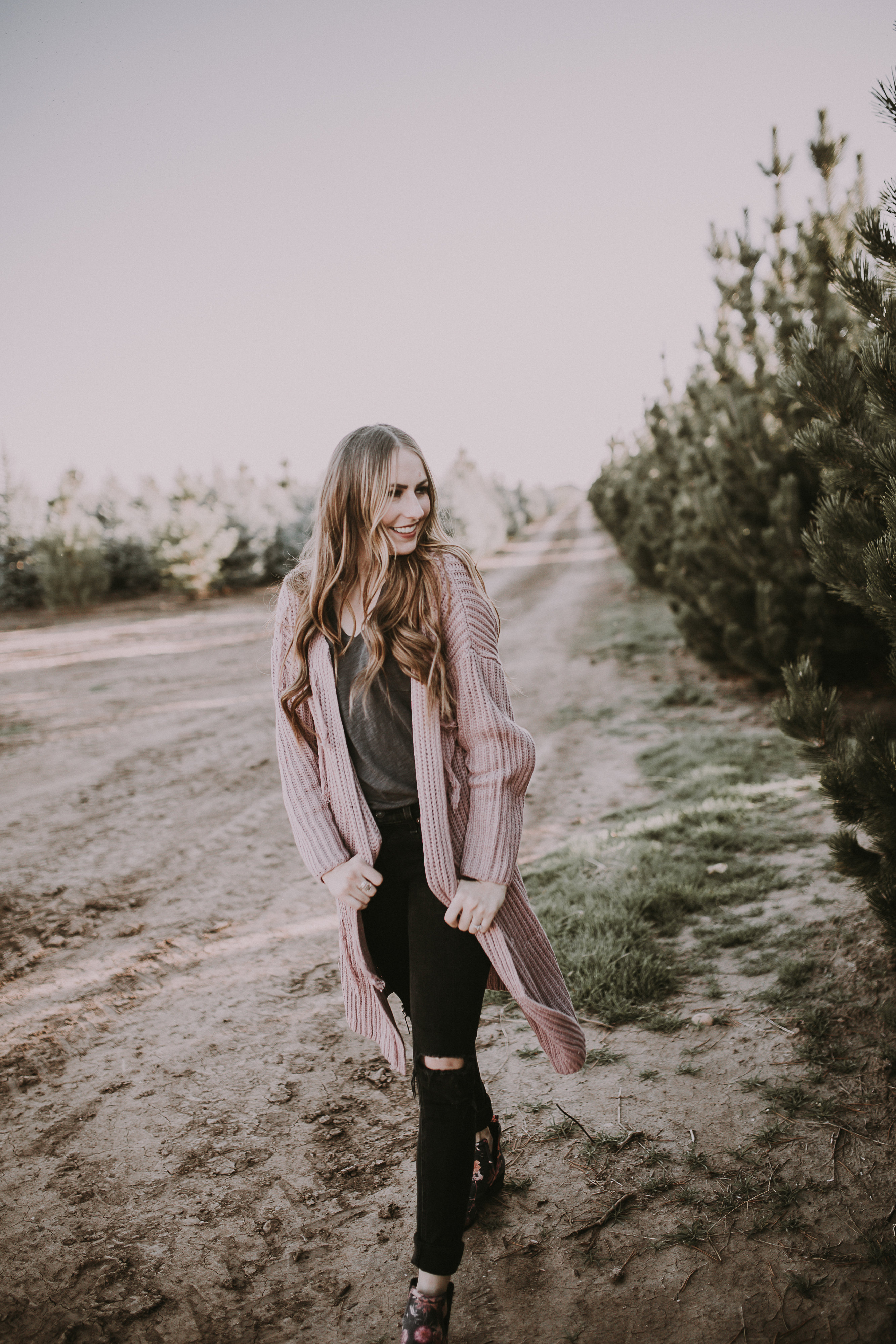 girl standing at tree farm in black jeans and light pink sweater with booties on