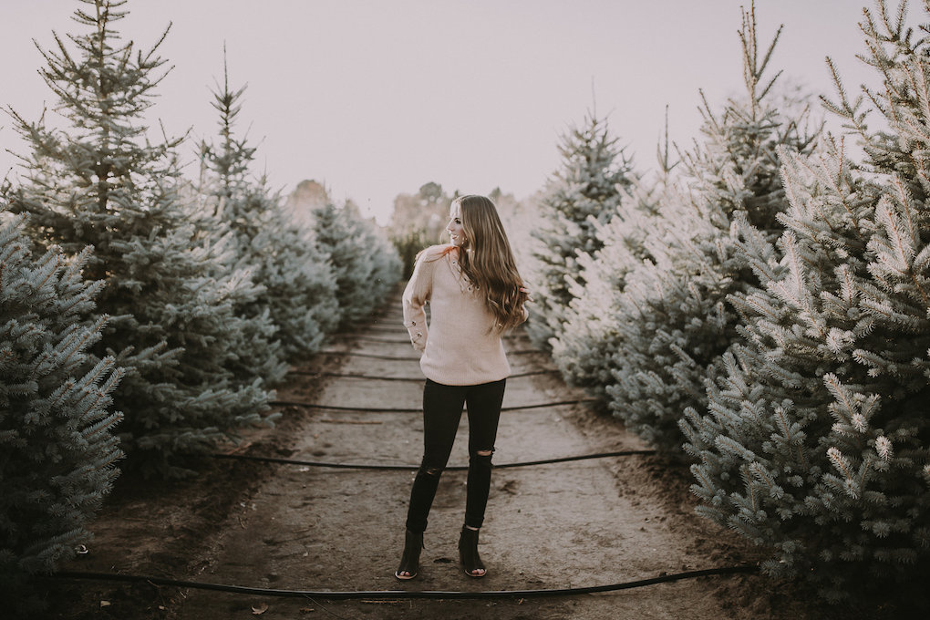 girl standing in tree farm in tan sweater with black jeans and peep toe toms booties