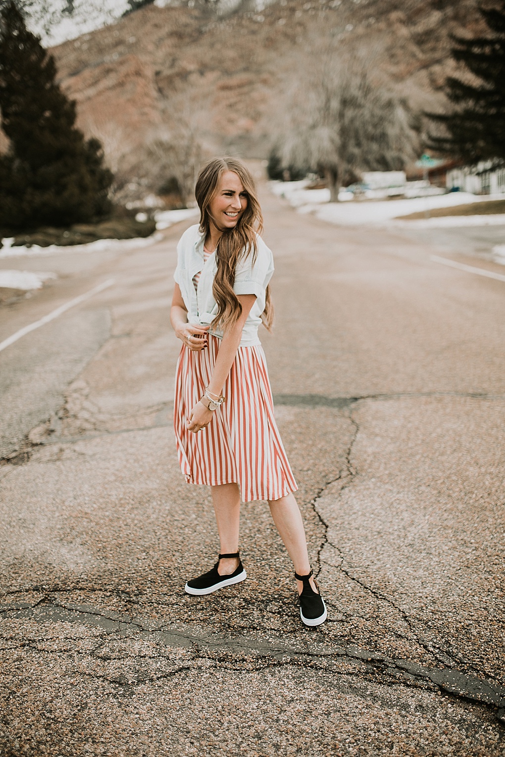 girl standing in road in pink stripe dress downeast basics with denim button up shirt over top with long loosely curled brown hair