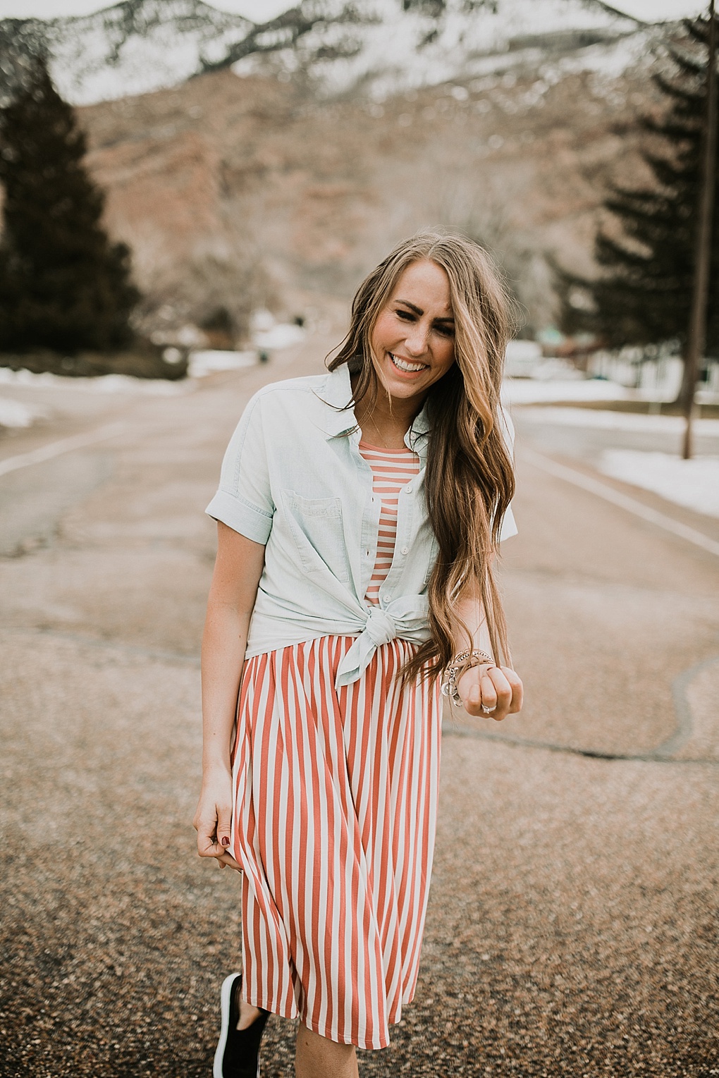 girl standing in road in pink stripe dress downeast basics with denim button up shirt over top with long loosely curled brown hair