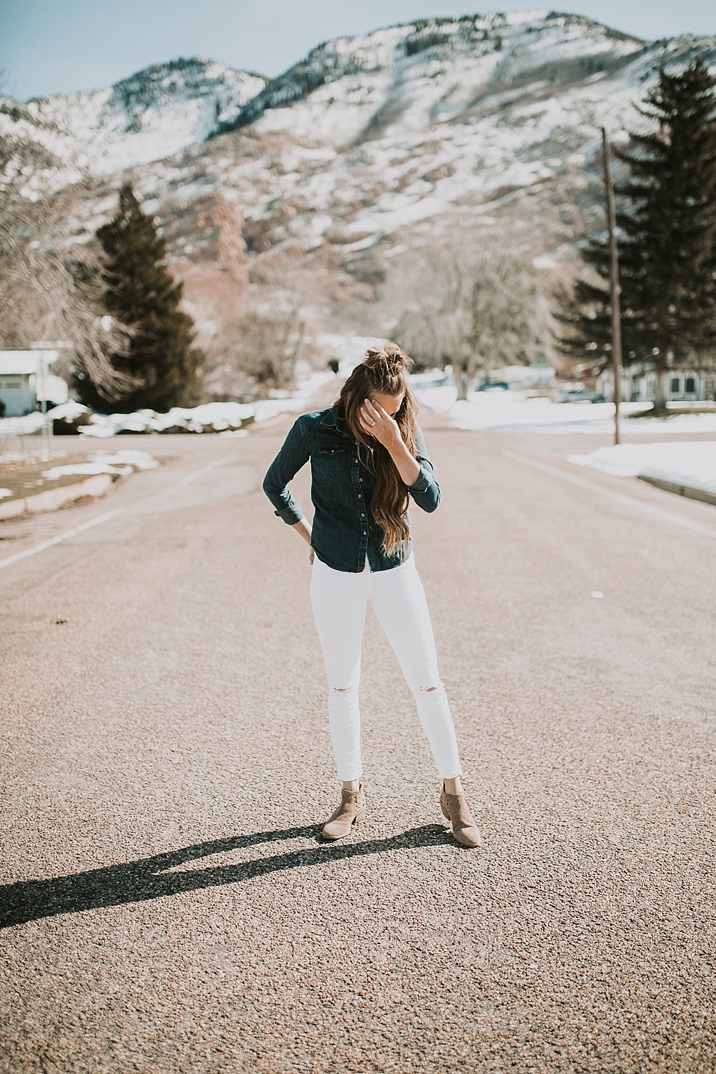 girl standing in the road in white jeans with a denim button shirt with loosely curled brown hair with half up top knot