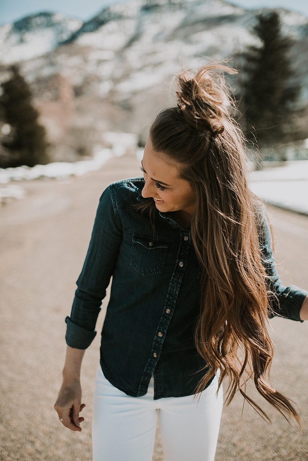 girl standing in the road in white jeans with a denim button shirt with loosely curled brown hair with half up top knot
