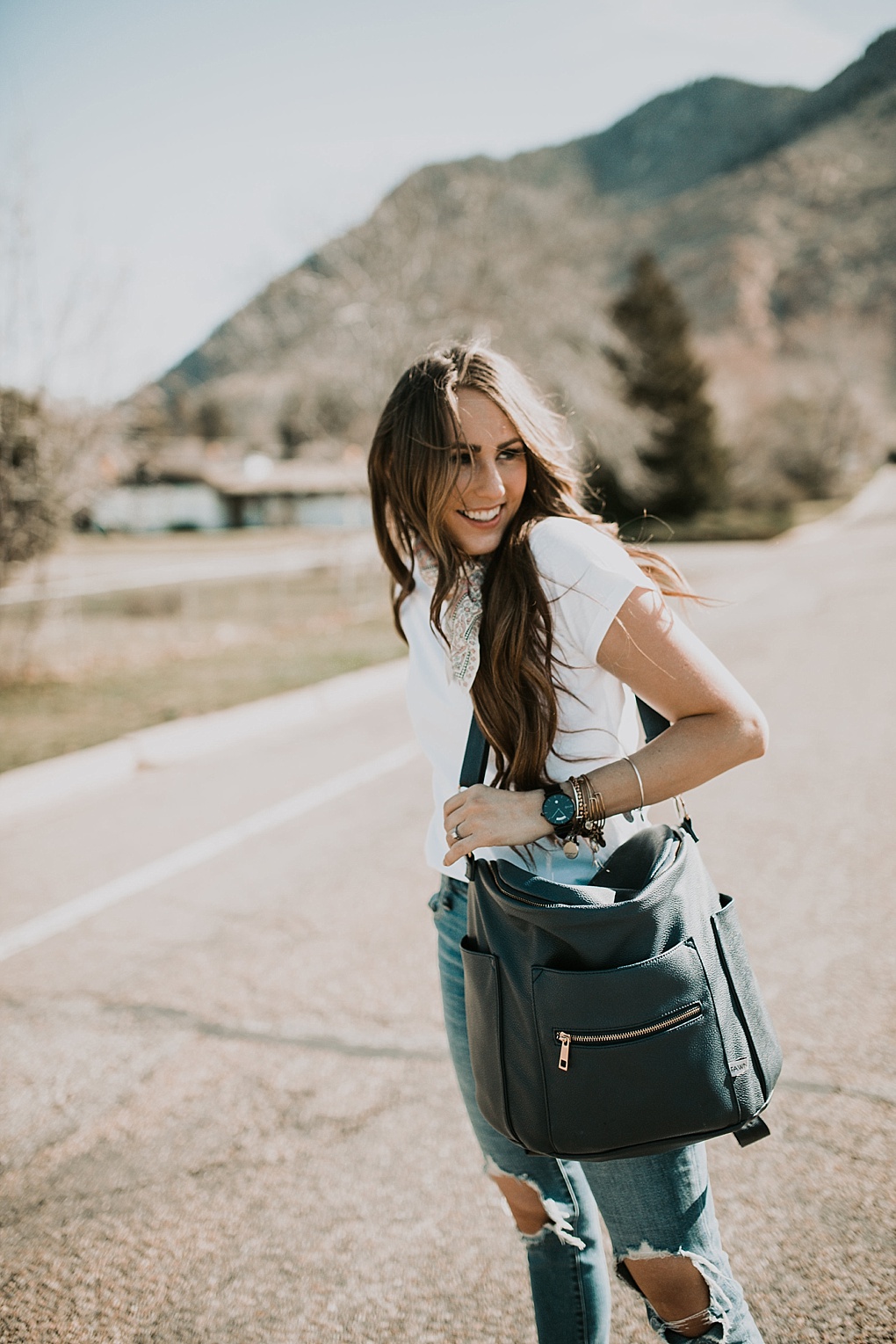 girl standing in road in white embroidered tee shirt with neck scarf and high waisted distressed denim jeans and fawn design diaper bag with long brown hair with caramel highlights loosely curled