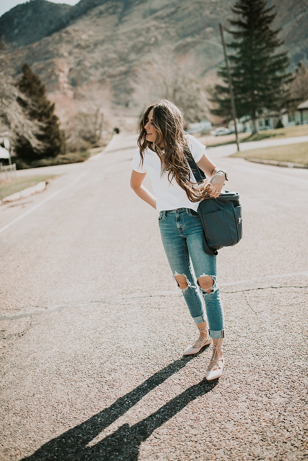 girl standing in road in white embroidered tee shirt with neck scarf and high waisted distressed denim jeans and fawn design diaper bag with long brown hair with caramel highlights loosely curled