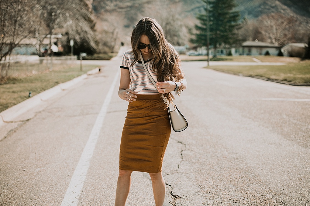 girl standing in street in navy and red stripe madewell tee shirt with gold suede pencil skirt and sneakers on with long loosely curled brown hair with caramel highlights michael kors crossbody bag and karen walker sunglasses