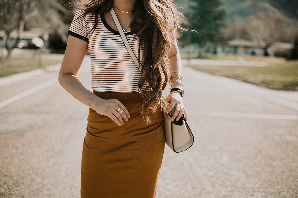 girl standing in street in navy and red stripe madewell tee shirt with gold suede pencil skirt and sneakers on with long loosely curled brown hair with caramel highlights michael kors crossbody bag and karen walker sunglasses