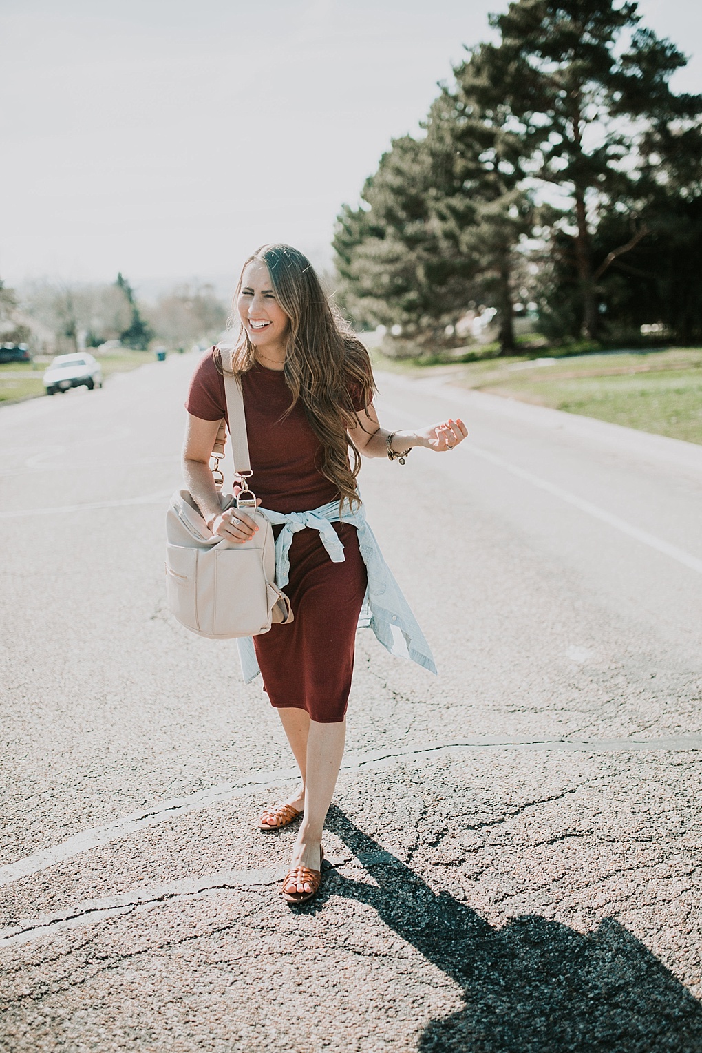 girl standing in road in maroon t shirt dress with target sandals and long loosely curled brown hair with caramel highlights with denim shirt tied around waist and fawn design bag