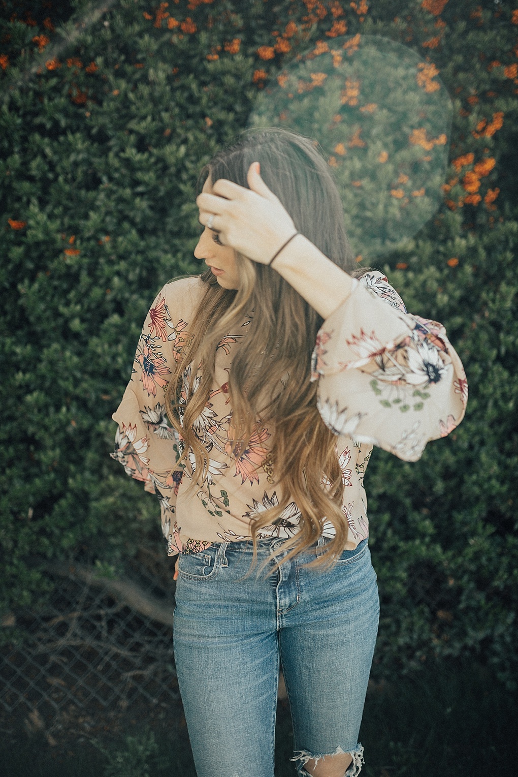 girl standing outside by greenery in floral ruffle top chicwish with long loosely curled brown hair with caramel highlights and high waisted levi jeans