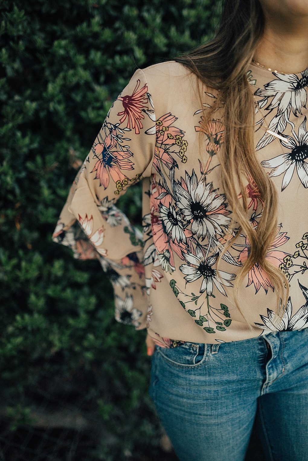 girl standing outside by greenery in floral ruffle top chicwish with long loosely curled brown hair with caramel highlights and high waisted levi jeans