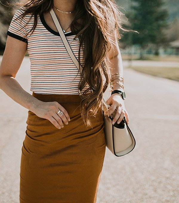 girl standing in street in navy and red stripe madewell tee shirt with gold suede pencil skirt and sneakers on with long loosely curled brown hair with caramel highlights michael kors crossbody bag and karen walker sunglasses