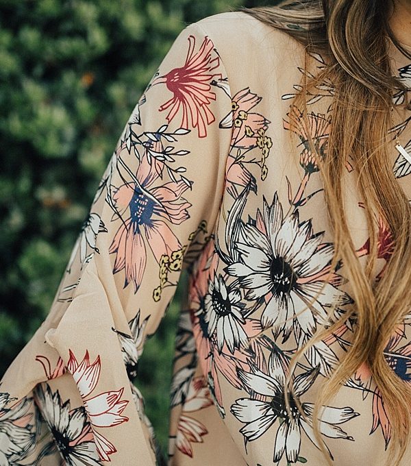 girl standing outside by greenery in floral ruffle top chicwish with long loosely curled brown hair with caramel highlights and high waisted levi jeans
